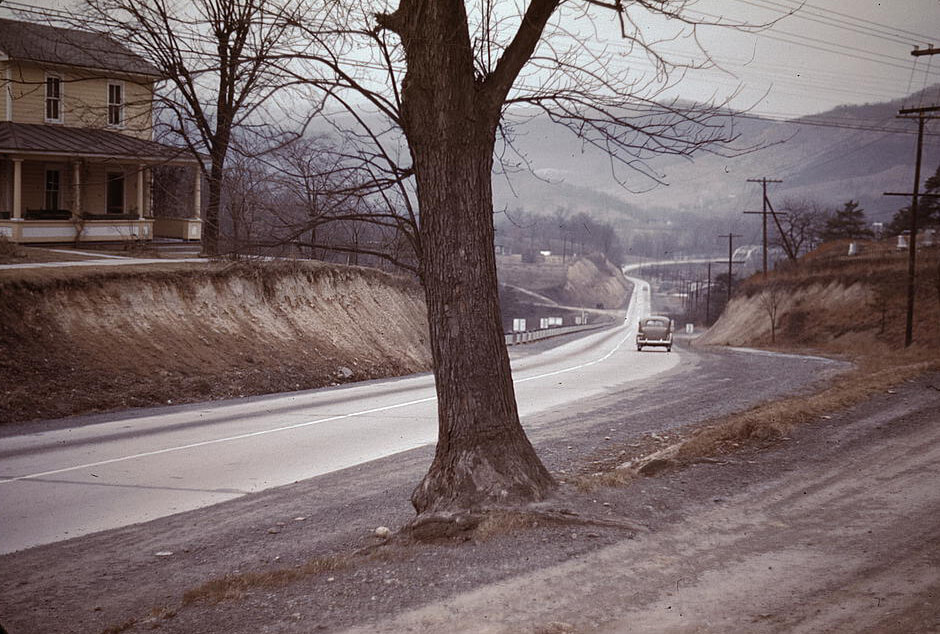 US 50 Looking West, Romney, West Virginia, 1942. Photograph originally published as part of the Farm Security Administration, Office of War Information Collection in the Library of Congress Prints and Photographs Division. Courtesy of Wikimedia Commons. Image is in public domain. A photograph with muted colors showing bare winter trees and a highway winding into the distance.
