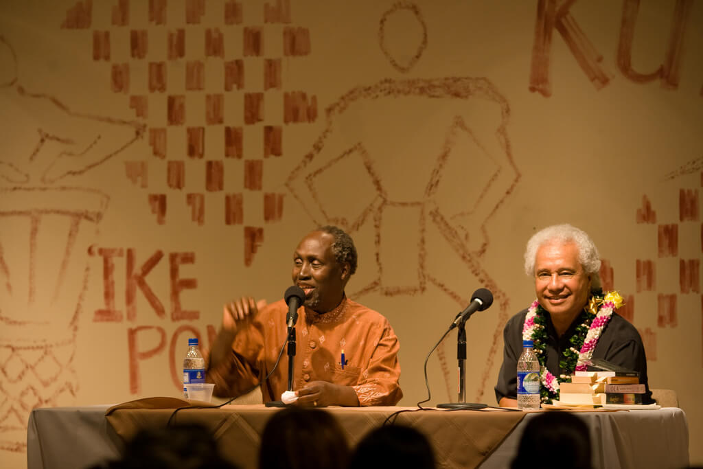 Samoan author Albert Wendt (right) with Kenyan writer Ngũgĩ wa Thiong'o (left), University of Hawaii at Manoa, Honolulu, Hawaii, April 30, 2008. Photograph by Flickr user Kanaka Rastamon. Creative Commons license CC BY-NC 2.0. Photograph of Samoan author Albert Wendt and Kenyan writer Ngũgĩ wa Thiong'o, sitting at a table. Ngũgĩ wa Thiong'o is speaking into a microphone and Wendt is smiling.