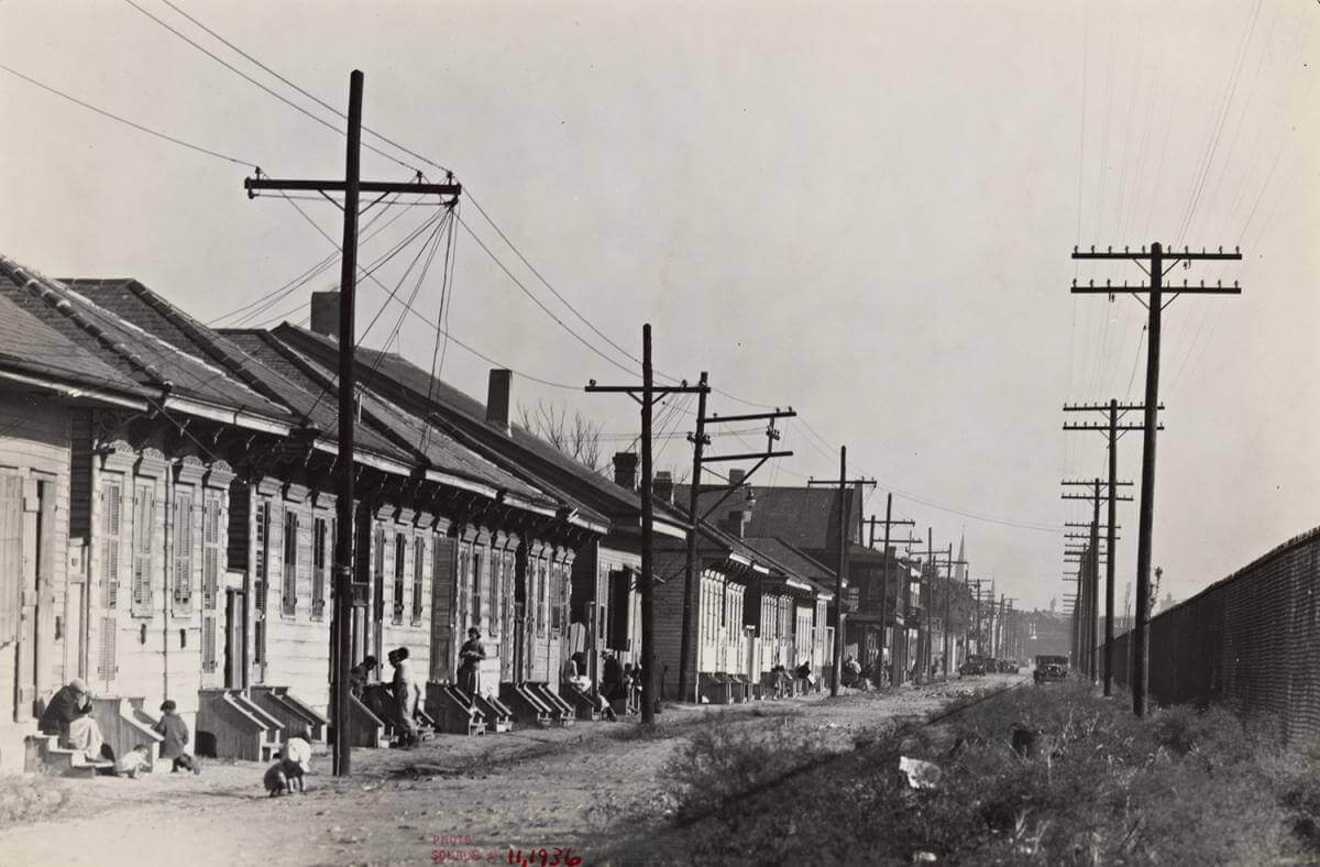 New Orleans "Negro" street, 1935. Photograph by Walker Evans. Courtesy of The New York Public Library Miriam and Ira D. Wallach Division of Art, Prints and Photographs, digitalcollections.nypl.org/items/d9317bb0-baca-0132-749d-58d385a7b928. Black and white photograph of a long line of white identical houses, with people and children sitting on the front steps.
