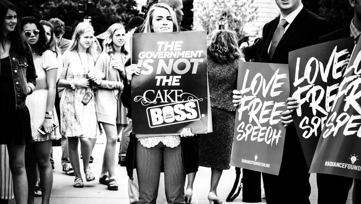 SCOTUS Rally, Masterpiece Cake Case, Washington, DC, June 4, 2018. Photograph by Flickr user Ted Eytan. Creative Commons license CC BY-SA 2.0. A black and white photograph of a crowd of protestors. A young woman holds a sign that says, "The government is not the cake boss." Other signs read, "Love free speech."