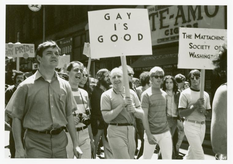 Black and white photograph of men and women marching with signs reading "Gay is good" and "Mattachine Society."