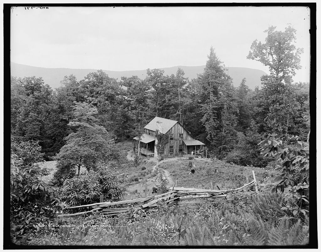 Black and white photograph of a wooden house in a clearing in the mountains behind an old wooden fence.